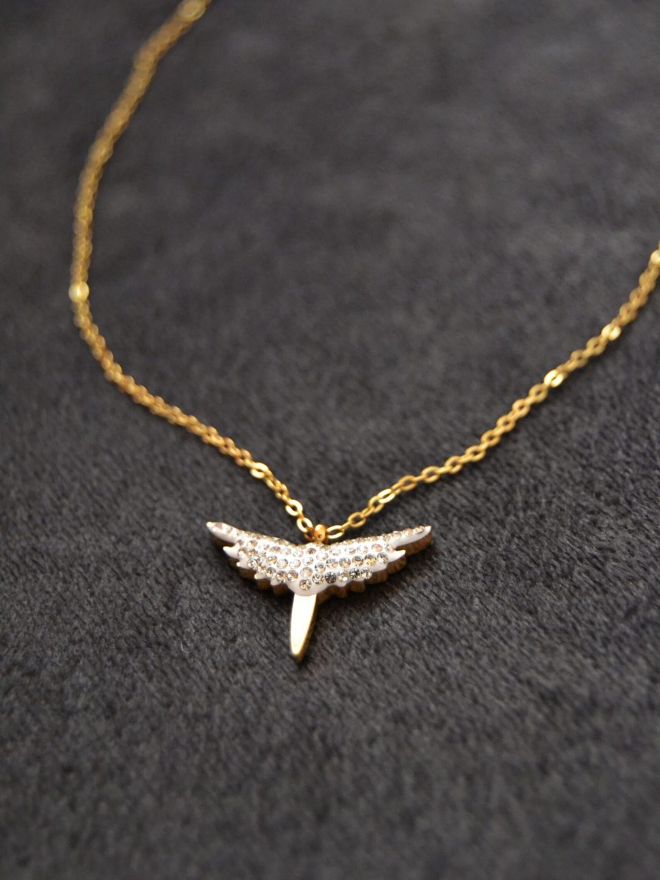 Close-up of a gold necklace featuring a diamond wing-shaped pendant on a dark background.
